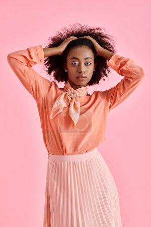 african american woman in peach blouse, midi skirt and neck scarf adjusting hair on pink backdrop
