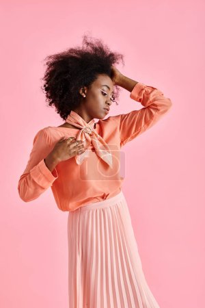 african american woman in peach blouse, midi skirt and scarf adjusting curly hair on pink backdrop