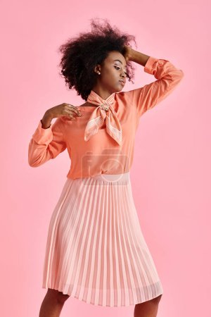 Photo for African american woman in peach blouse, midi skirt and scarf adjusting curly hair on pink backdrop - Royalty Free Image