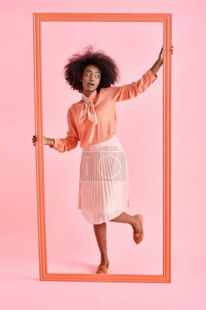 curly african american woman in peach blouse and midi skirt strikes pose near frame on pink backdrop