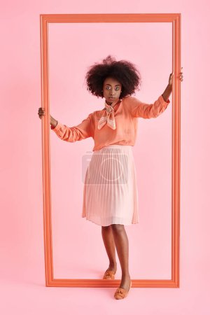 Foto de Joven africana americana mujer en melocotón blusa y midi falda huelgas pose cerca marco en rosa telón de fondo - Imagen libre de derechos
