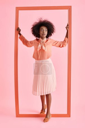 young african american woman in peach blouse and midi skirt strikes pose near frame on pastel pink