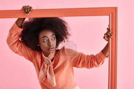 Foto de Joven afroamericana mujer en melocotón pelusa blusa y cuello bufanda posando en marco sobre rosa telón de fondo - Imagen libre de derechos