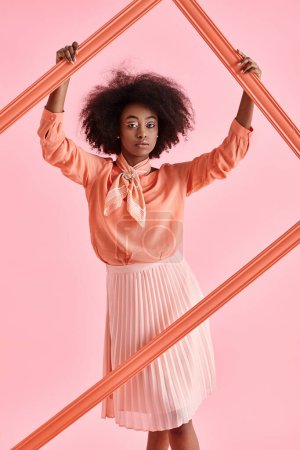 bonita mujer afroamericana en blusa de pelusa de melocotón y falda midi posando en marco sobre fondo rosa