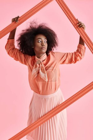 Foto de Mujer afroamericana soñadora en blusa de pelusa de melocotón y falda midi posando en marco sobre fondo rosa - Imagen libre de derechos