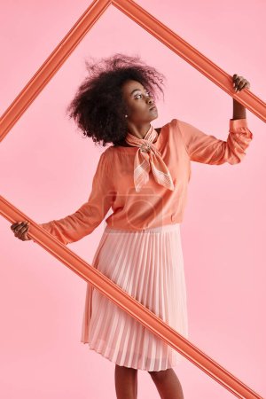 dreamy african american girl in peach fuzz blouse and midi skirt posing in frame on pink backdrop