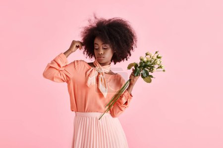 african american woman in peach fuzz attire holding flowers and looking down on pink background