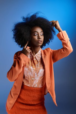 lockige afrikanisch-amerikanische Frau in pfirsichfarbener Flauschkleidung mit Blazer, der vor blauem Hintergrund wegschaut