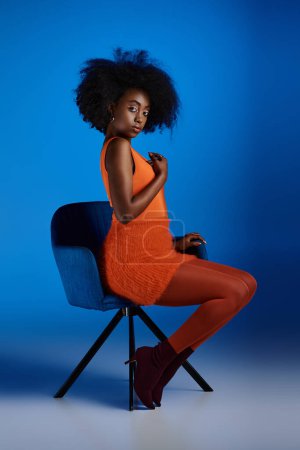 Foto de Elegante modelo afroamericano en vestido texturizado y tacones altos sentado en silla sobre fondo azul - Imagen libre de derechos
