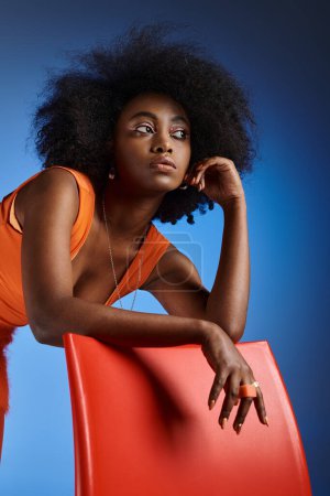 Photo for Dreamy african american woman in peach fuzz dress posing next to chair on vivid blue background - Royalty Free Image