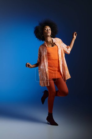 Schicker Look eines afrikanisch-amerikanischen Models in gemustertem Hemd und orangefarbenem Kleid, das auf blauem Hintergrund posiert