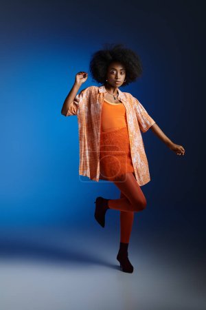 Schicker Look einer Afroamerikanerin in gemustertem Hemd und orangefarbenem Kleid, die auf blauem Hintergrund posiert