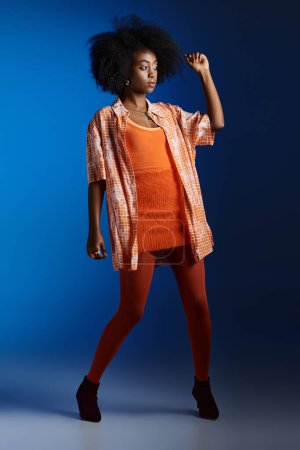 stylisches Aussehen eines afrikanisch-amerikanischen Models in gemustertem Hemd und orangefarbenem Kleid, das vor blauem Hintergrund posiert