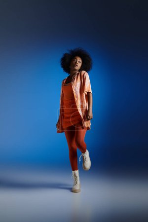 trendy look of african american model in patterned shirt and orange dress posing on blue backdrop