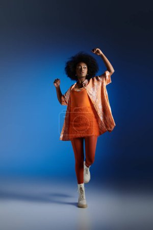 trendy look of african american model in patterned shirt and textured dress posing on blue backdrop