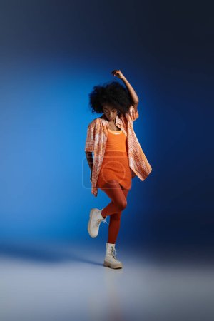 Téléchargez les photos : Modèle afro-américain élégant en chemise à motifs et robe texturée posant sur fond bleu - en image libre de droit