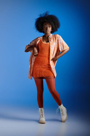 fashionable african american woman in patterned shirt and textured dress posing on blue backdrop