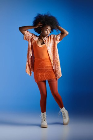 fashionable african american model in patterned shirt and trendy dress posing on blue backdrop