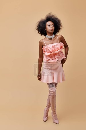 brünette junge afrikanisch-amerikanische Frau in pfirsichfarbenem Rüschenoberteil und Overknee-Stiefeln auf beigem Hintergrund