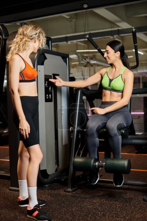 vigorous female coach consulting her blonde cheerful client in gym near shoulder press machine