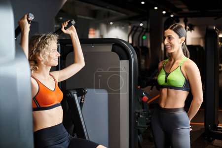 energische weibliche Trainerin berät ihren blonden freudigen Kunden im Fitnessstudio in der Nähe der Schulterpresse