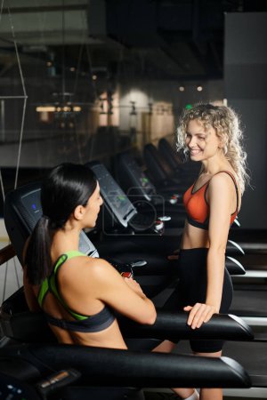 positive appealing female coach and her client in sportwear exercising actively on treadmill in gym