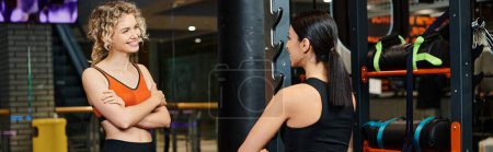 Téléchargez les photos : Entraîneur féminin joyeux aidant son client joyeux blonde à étirer ses muscles tout en salle de gym, bannière - en image libre de droit