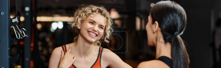 Téléchargez les photos : Entraîneur féminin joyeux aidant son client joyeux blonde à étirer ses muscles tout en salle de gym, bannière - en image libre de droit