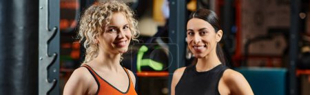 Foto de Alegre atractivo entrenador femenino y su cliente en ropa deportiva sonriendo a la cámara en el gimnasio, pancarta - Imagen libre de derechos
