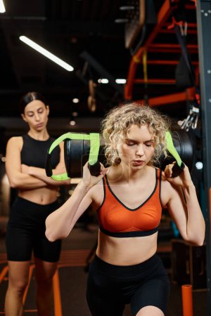 focus on blonde attractive woman exercising with power bag with her blurred female coach on backdrop