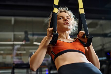 good looking blonde woman in comfortable sportwear exercising with pull ups equipment while in gym