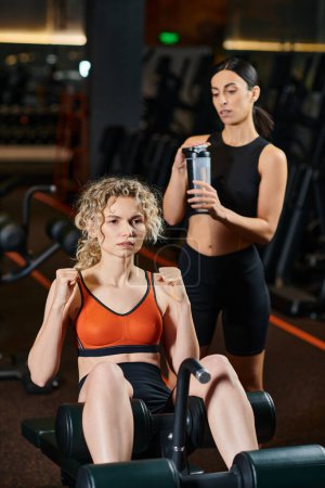 attractive athletic female coach consulting blonde beautiful woman how to pump press while in gym