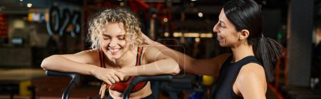 Photo for Cheerful female coach helping her beautiful client to use cross trainer in gym, water bottle, banner - Royalty Free Image