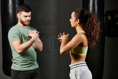 A male trainer guides a woman in self-defense training, standing together in front of a punching bag at the gym.