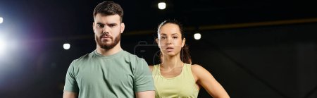 Un entrenador masculino y una mujer en un gimnasio, ambos de pie con confianza uno al lado del otro.