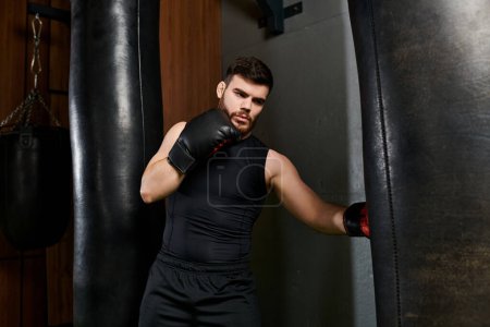 A handsome man with a beard, wearing a black tank top and boxing gloves, practices his punches on a heavy bag in the gym.