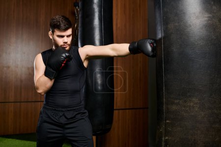 Un hombre guapo con barba y camiseta negra y guantes de boxeo golpea una bolsa en el gimnasio.
