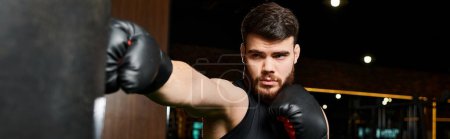 Photo for A handsome man with a beard, wearing boxing gloves, fiercely punches a punching bag in a gym. - Royalty Free Image