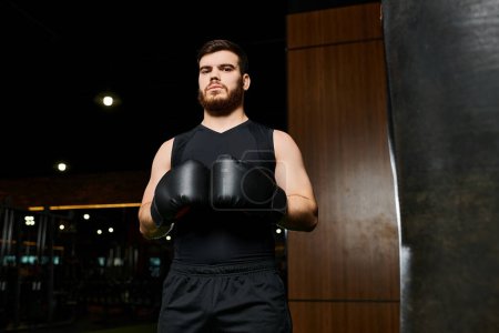 A bearded, handsome man wearing boxing gloves stands confidently next to a punching bag in a gym.