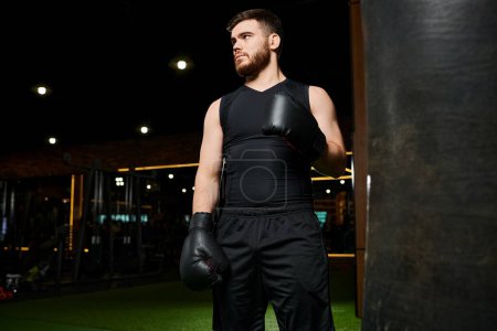 Un hombre barbudo con una camiseta negra lanza puñetazos a un saco de boxeo colgante en un gimnasio.