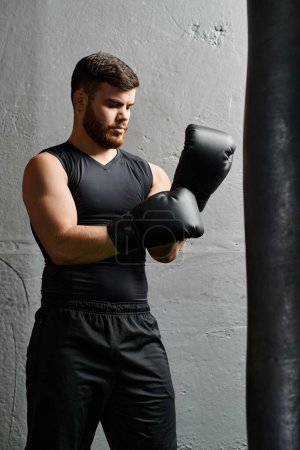 Téléchargez les photos : Un bel homme avec une barbe se tient à côté d'un sac de boxe dans un gymnase, pratiquant des techniques de boxe avec concentration et détermination. - en image libre de droit