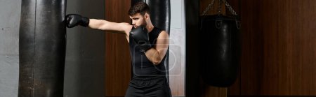 Ein gutaussehender Mann mit Bart trägt ein schwarzes Hemd und schwarze Handschuhe und boxt in einem Ring mit einem Boxsack in einer Turnhalle.