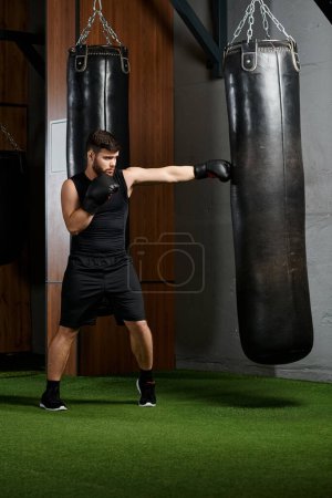 Hombre barbudo guapo con camisa negra y pantalones cortos golpea vigorosamente un saco de boxeo en un entorno de gimnasio.