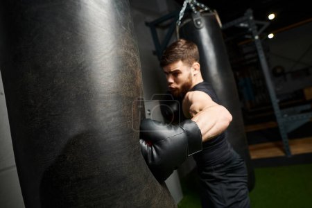 A handsome man with a beard wearing a black shirt and boxing gloves, focuses on punching a bag in a gym.