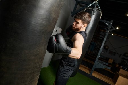 Hombre guapo con barba en camiseta negra y guantes de boxeo lanzando puñetazos a un saco de boxeo en un gimnasio.