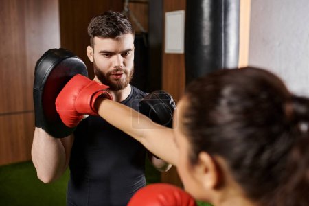 A male trainer assists a brunette sportswoman, both dressed in active wear, as they engage in a boxing session in a gym.