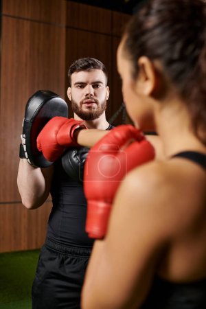 A man in red boxing gloves coaches a brunette sportswoman in active wear as they practice boxing in a gym.