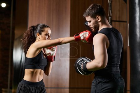Un entrenador masculino guía a una deportista morena en ropa activa mientras practican boxeo dentro de un gimnasio.