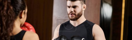 A male trainer in boxing gloves talking to a brunette sportswoman in active wear in a gym setting.