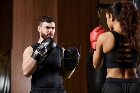 A male trainer guides a brunette sportswoman in active wear through a boxing session in a gym.